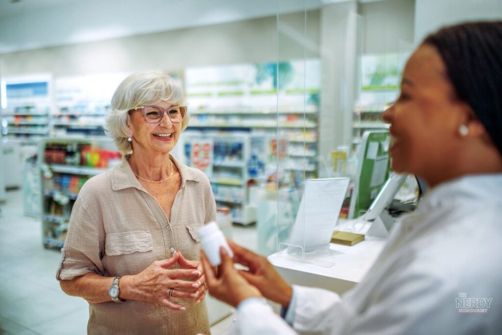 Elderly American woman smiling at a pharmacy counter as a pharmacist hands her a prescription bottle — among the seniors covered by the Trump Medicare $90 payment for seniors