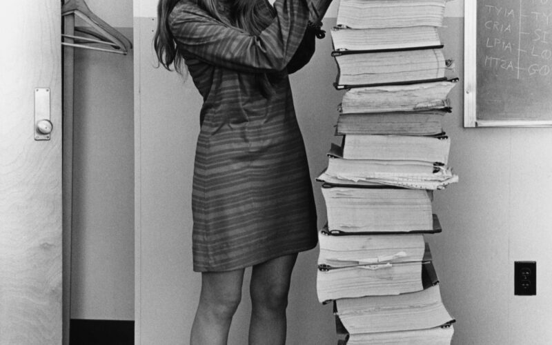 Margaret Hamilton stands smiling beside a towering stack of printed Apollo Guidance Computer program listings that she and her MIT team developed, in the famous 1969 Draper Laboratory photograph.
