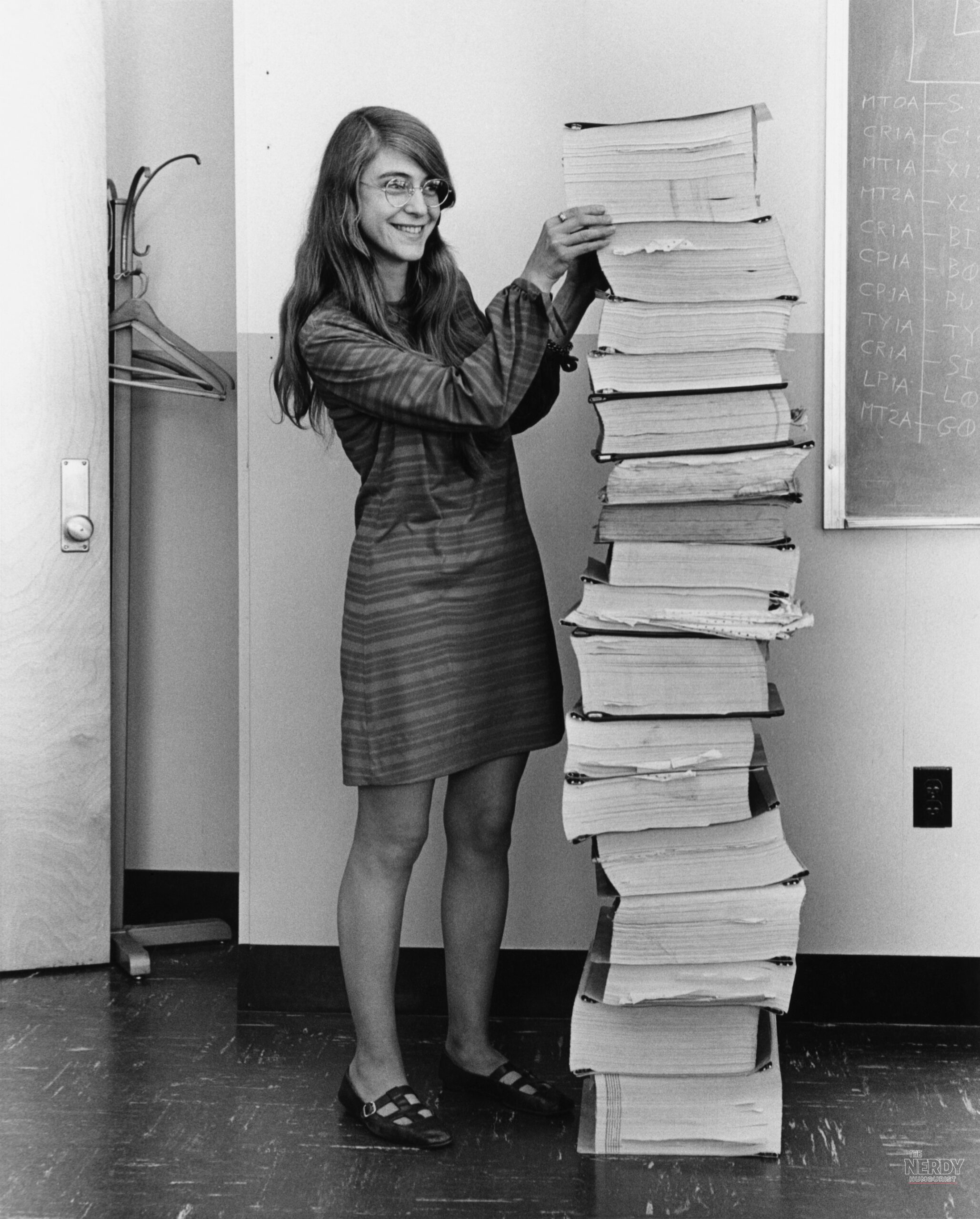 Margaret Hamilton stands smiling beside a towering stack of printed Apollo Guidance Computer program listings that she and her MIT team developed, in the famous 1969 Draper Laboratory photograph.