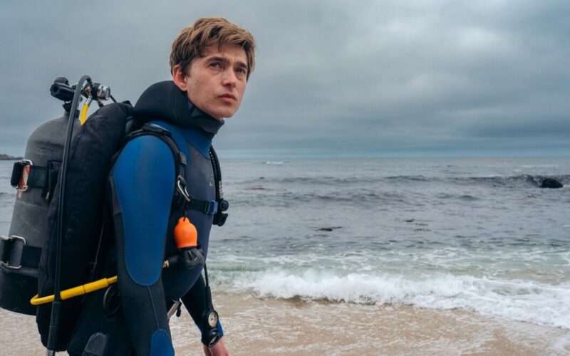 Austin Abrams as Jay Gardiner in a wetsuit with scuba gear, walking along the beach in the Whalefall movie